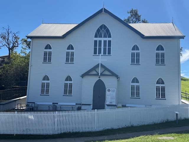 The Bulimba Uniting Church building today — white weatherboard with a tall central gothic window, steep gable, and the surrounding white picket fence under a blue Queensland sky.